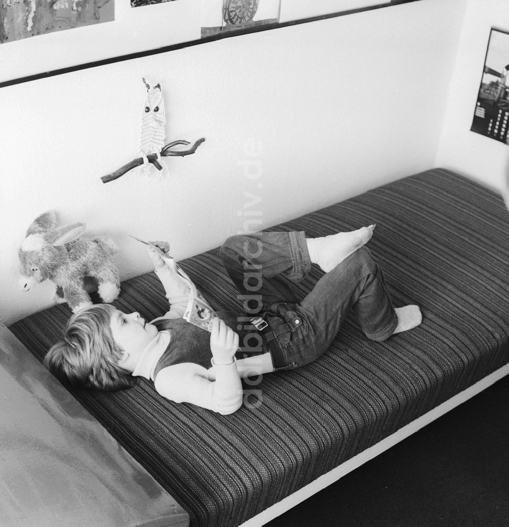 DDR-Bildarchiv: Berlin - Ein Kind liegt auf einer Liege in seinem Kinderzimmer und liest in Berlin, der ehemaligen Hauptstadt der DDR, Deutsche Demokratische Republik