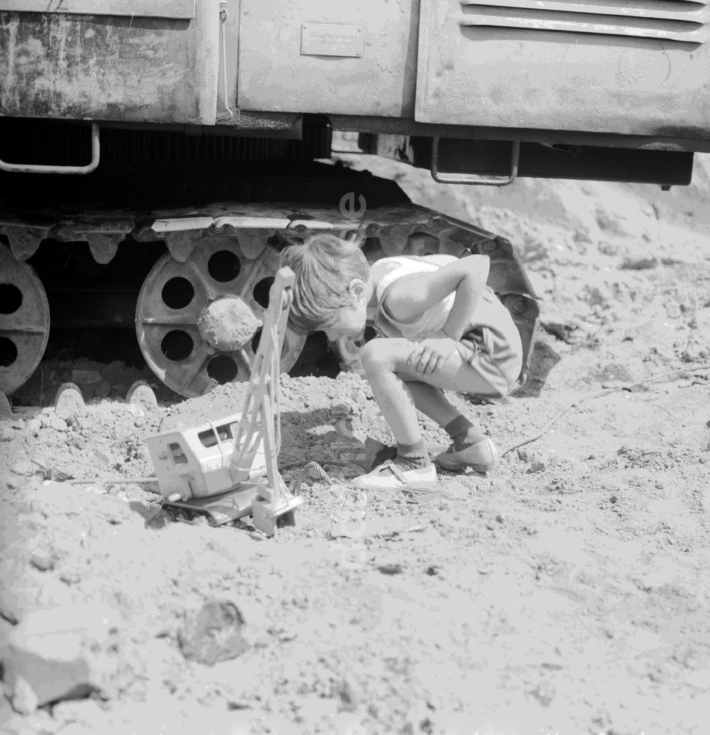 DDR-Fotoarchiv: Berlin - Kleiner Junge in Lederhosen spielt mit seinem Holzbagger auf einer Baustelle neben einem Großbagger in Berlin, der ehemaligen Hauptstadt der DDR, Deutsche Demokratische Republik