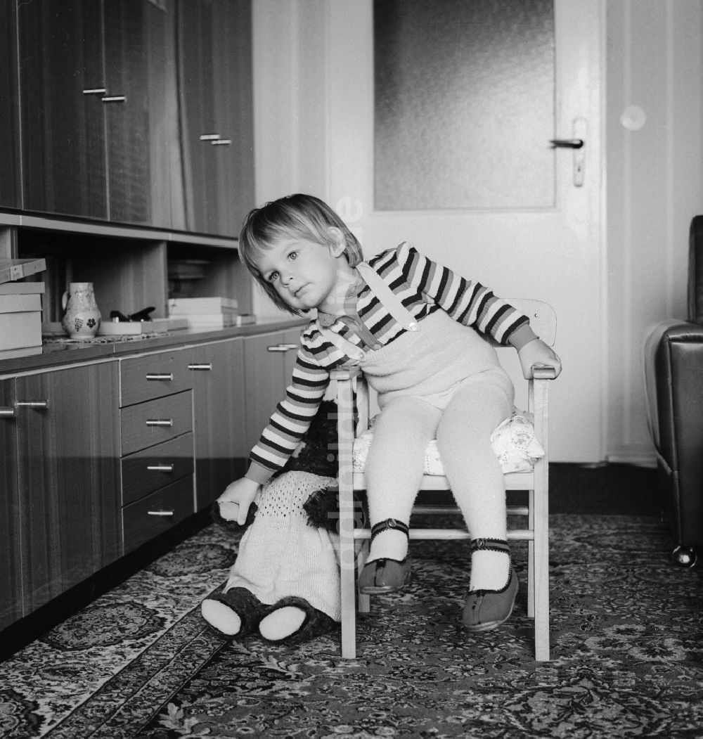 DDR-Fotoarchiv: Berlin - Kleines Kind sitzt verträumt auf einem Stuhl und greift nach seinem Spielzeug in Berlin