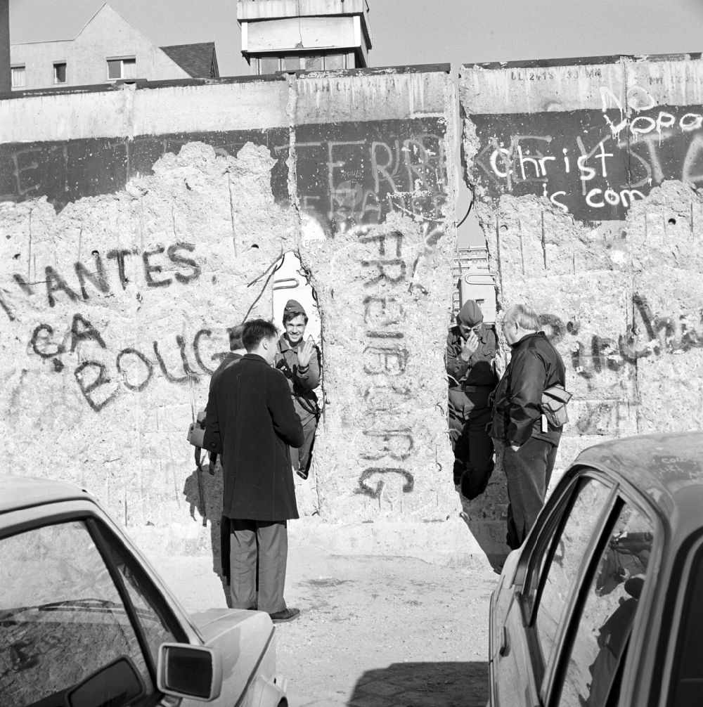 Berlin: Zwei Soldaten der Grenztruppen der DDR schauen durch ein Loch in der Berliner Mauer und unterhalten sich mit Passanten in Berlin